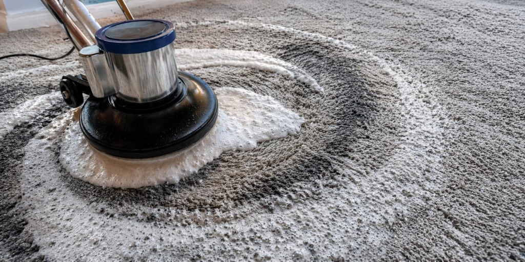 Máquina profesional rotativa limpiando moquette con espuma sobre alfombra gris